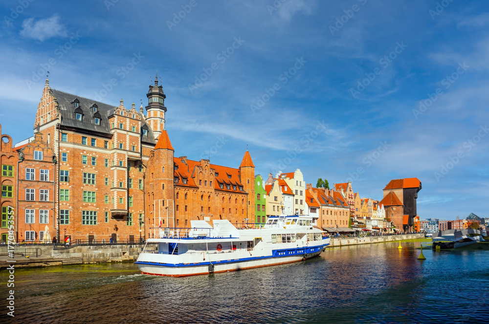 Fototapeta premium Scenic view of two boats navigating the Motlawa River alongside historic colorful buildings in Gdansk, Poland. Bright blue skies enhance this vibrant European cityscape