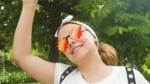 Smiling young woman enjoying a sunny day outdoors, holding a coffee cup high while wearing a white bandana and unique orange paper glasses, embodying fun, joy, and summer vibes