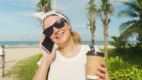 Young woman smiling while having a phone call and holding a coffee cup on a sunny tropical beach boardwalk, enjoying her summer vacation near the ocean with palm trees