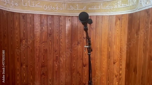 A peaceful wooden prayer corner inside an empty mosque, with a microphone stand prepared for the imam’s sermon, soft patterned carpet, serene lighting capturing the calm atmosphere of Islamic worship