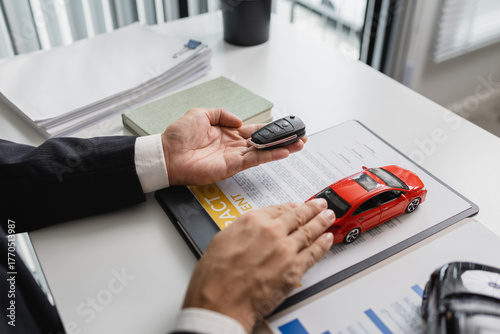 A businessman in a suit is presenting car insurance and contract documents with miniature car models on the desk, symbolizing vehicle insurance, financial planning, and legal agreements.