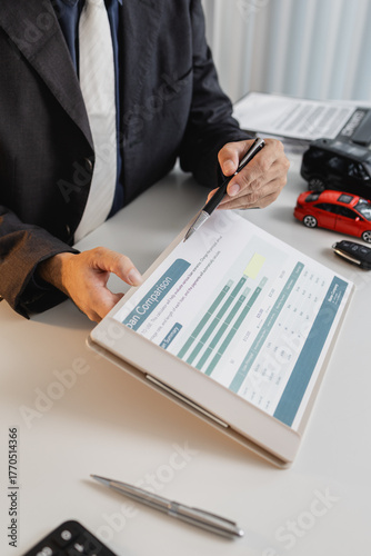 A businessman in a suit is calculating loan details, reviewing financial documents, and holding car keys near a calculator and toy car models, representing auto loan and finance services.