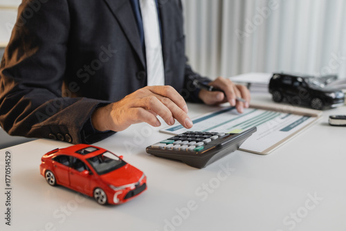 A businessman in a suit is calculating car loan options using a calculator and comparison chart, with model cars and car keys on the desk, symbolizing finance, leasing, and investment planning.