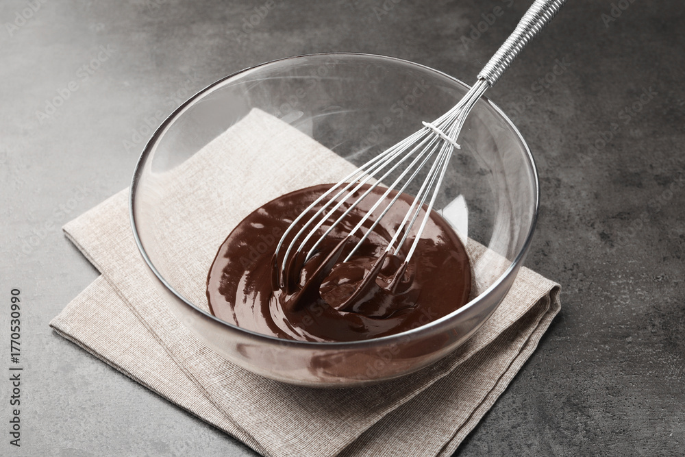 Fototapeta premium Bowl with melted chocolate and whisk on grey table, closeup