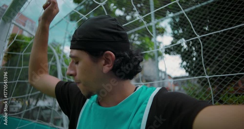 Hispanic soccer player holds goal net in thoughtful silence after match, looking away as he reflects on life, choices, and personal meaning on quiet urban field
