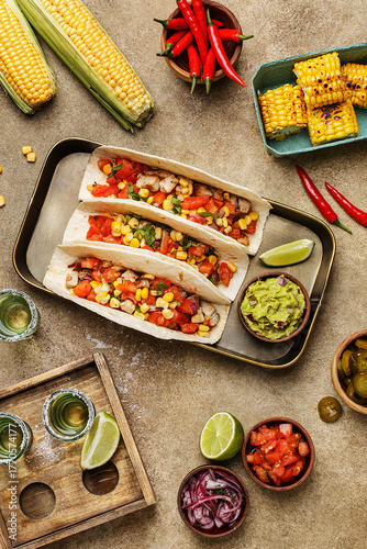 Tacos with chicken, tomatoes and corn with guacamole sauce, lime, corn cobs, chili peppers and tequila shots on beige concrete background. Overhead view, flat lay. Mexican party snacks