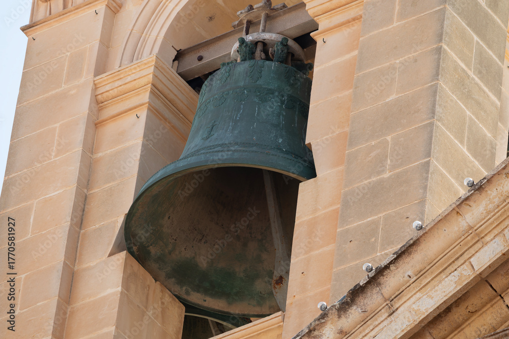 Obraz premium A large, ancient-looking bell with green patina hangs within a stone bell tower, featuring intricate carvings on its upper rim.