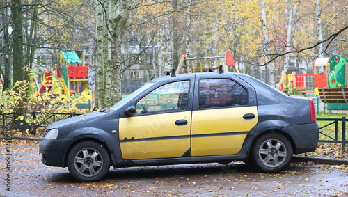 A dark gray car with yellow doors is parked near a playground fence, Podvoyskogo Street, Saint Petersburg, Russia, October 28, 2025