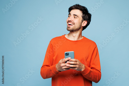Young fun Caucasian man he wear orange sweatshirt casual clothes hold in hand use mobile cell phone look aside isolated on plain pastel light blue cyan background studio portrait. Lifestyle concept