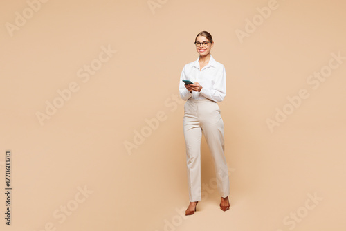 Full body young smiling successful employee business woman corporate lawyer wear classic formal white shirt work in office hold in hand use mobile cell phone isolated on plain beige background studio