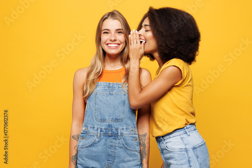 Two young friends buddies women wear denim casual clothes together whispering gossip and tells secret behind her hand sharing news isolated on plain yellow orange background studio. Lifestyle concept.