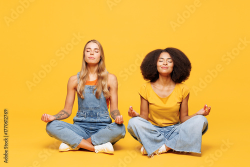 Full body two young friends buddies women wear denim casual clothes sit together hold spreading hands in yoga om aum gesture relax meditate try to calm down isolated on plain yellow orange background.