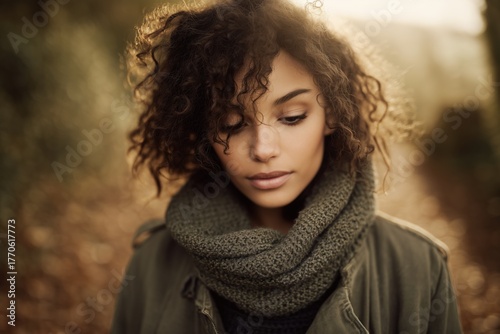 Young woman with curly hair wearing scarf, autumn background, looking down, soft focus