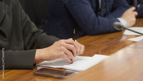 Close-up of a lawyer or official in a suit holding a pen over documents at a wooden desk during a meeting; smartphone and mic visible, focus on hands and paperwork. Photo