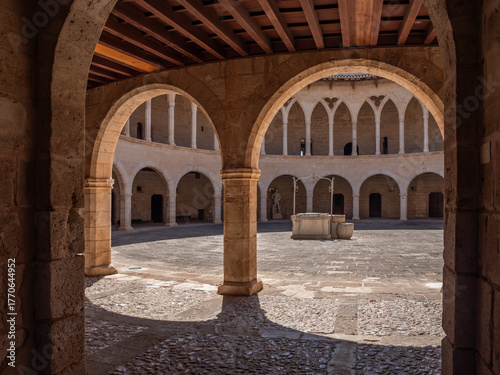 Castell de Bellver. Palma de Mallorca, historic courtyard features a circular design with stone arches and a central fountain, bathed in sunlight, Majorca