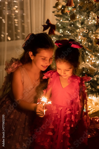 two little girls are watching a sparkler near a Christmas tree