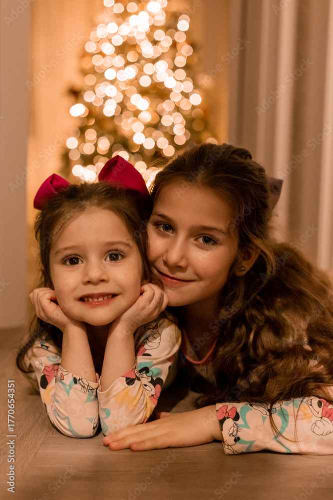 Fototapeta premium two little girls in pajamas by the Christmas tree at home on Christmas night