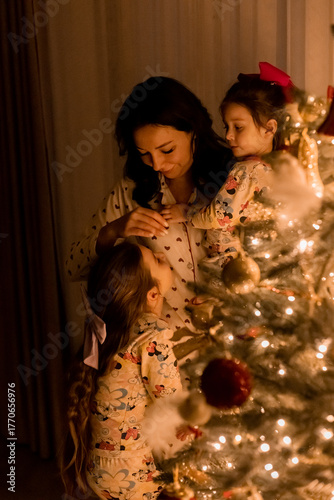 A mother and two little daughters in pajamas decorate a Christmas tree at home on New Year's Eve