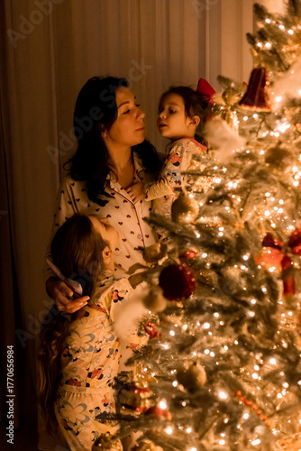 A mother and two little daughters in pajamas decorate a Christmas tree at home on New Year's Eve