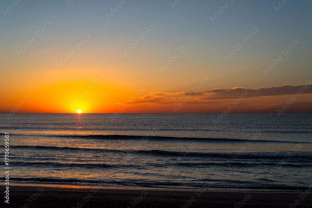 Obraz premium colorful sunset paints sky with shades of orange and yellow as it sets over calm ocean waves. tranquil beach scene invites relaxation and reflection during serene evening