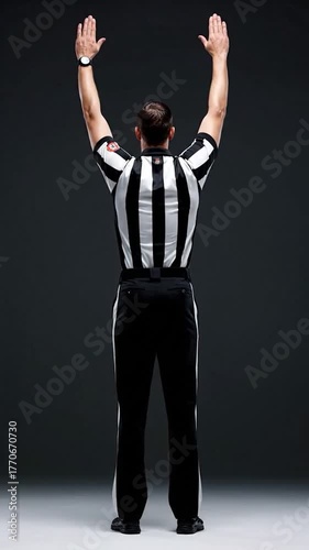 A referee raises arms in the air, signaling a call, viewed from the back. Black and white uniform