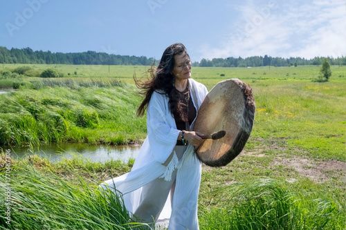 Beautiful female shaman in a trance in the white dress drumming in the natural environment