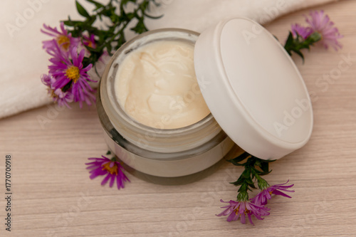 A glass container for cosmetic cream with lilac flowers. On a wooden background.