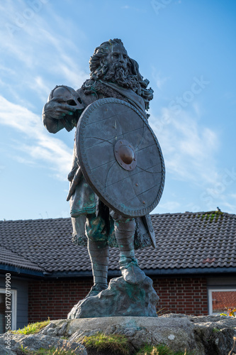 King Harald, Fairhair statue, bronze statue of a bearded man holding a shield stands on a rocky pedestal in front of a brick building under a clear blue sky, Haugesund, Norway