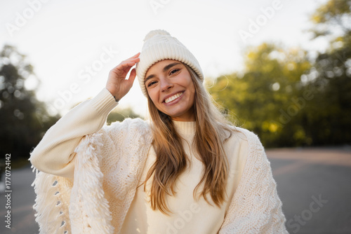 attractive smiling young blond woman walking in winter park having fun in warm white knitted sweater, cape and hat, cold season