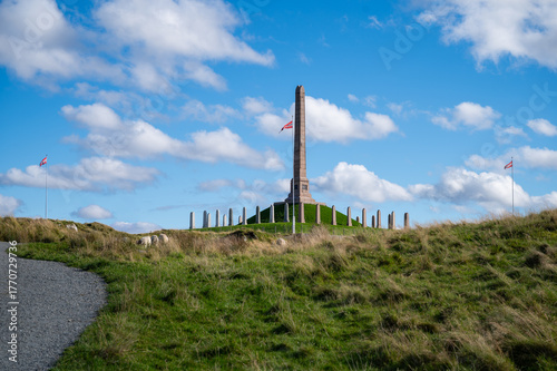 Haraldshaugen, Haugesund, Norway, tall monument stands on a grassy hill, surrounded by evenly spaced pillars, with sheep grazing nearby and two flags flying on poles