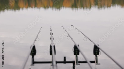 Fishing rods on tripod pointed toward calm lake, minimalist autumn landscape reflection on water, peaceful outdoor recreation and patience concept in nature, shallow depth of field