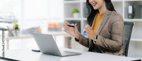 Digital Dialogue: A woman in a smart casual business attire is engaged in a video conference, appearing engaged, at a modern desk with a laptop and organized surroundings.