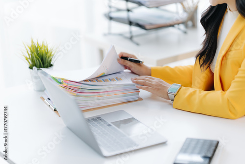 Analysing Paperwork: A focused individual in a yellow blazer meticulously reviews a stack of documents at a clean office desk, surrounded by a laptop, pen, and a touch of greenery.