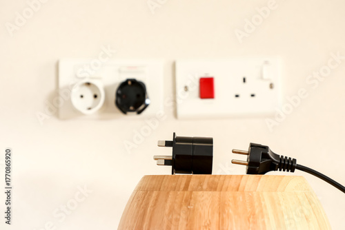 An electrical plug and a black travel adapter on a wooden surface, with blurred power sockets in the background. Minimalist image representing technology, connectivity, and modern travel.