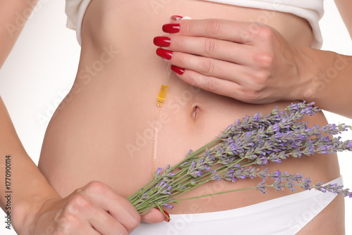 Close-up of a woman applying cosmetic oil with a dropper on her belly near the navel while holding lavender flowers, studio shot on a white background.