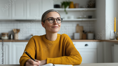 Woman writing in a notebook, wearing glasses and a yellow sweater, smiling while looking away with a thoughtful and enthusiastic expression, planning her day and future success in a kitchen setting