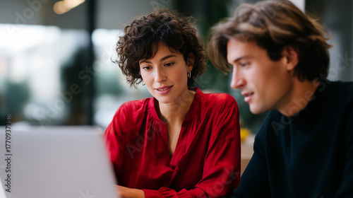 two colleagues collaborating at cafe table with laptop, woman in red blouse pointing at screen while man listens, modern workspace atmosphere, teamwork and communication concept, n