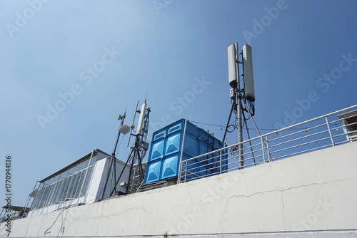 antennas and signal transmitting devices on the roof of a white building with a blue water tank next to it, the clear sky in the background emphasizes the elements of modern technology
