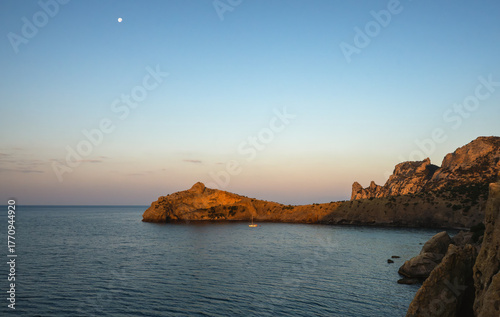 Yacht on calm sea near rocky coast at sunset