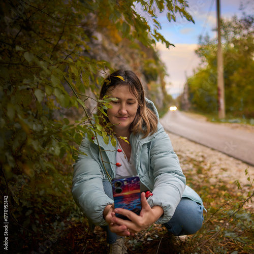 A woman takes a selfie on the street for social media. She squats near roadside trees.