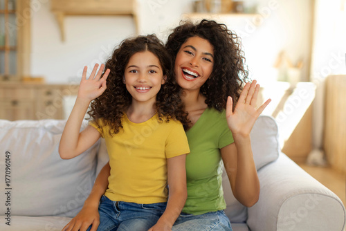 Hello. Happy mom and her preteen daughter waving hands and smiling at camera, sitting on sofa at home. Distance communication concept