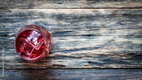 Top view of a refreshing red drink with ice cubes served in a clear glass on rustic wooden table.