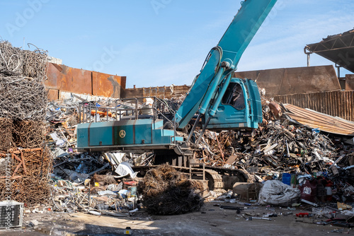 Excavator loading scrap metal onto truck in junkyard. Industrial excavator working at metal recycling yard. Excavator handling metal waste at recycling facility.