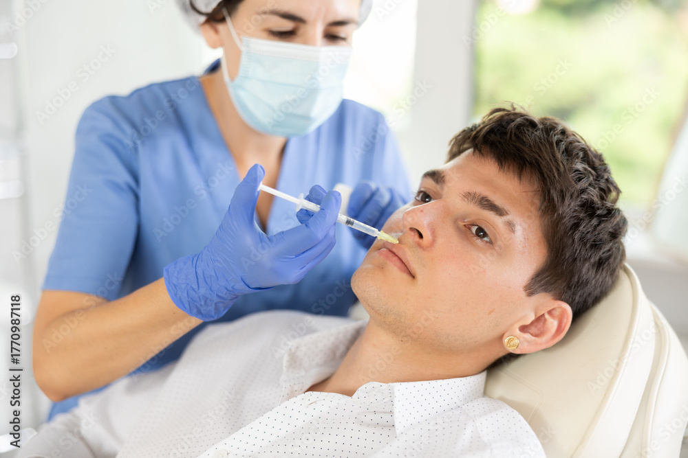 Fototapeta premium Young male patient lying on clinical chair undergoing face care procedure by injection method