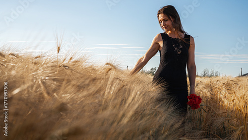 Elegant woman in a black dress in a golden wheat field at sunset, holding red flowers. Evokes rustic beauty, tranquility, and natural grace. Summer romance