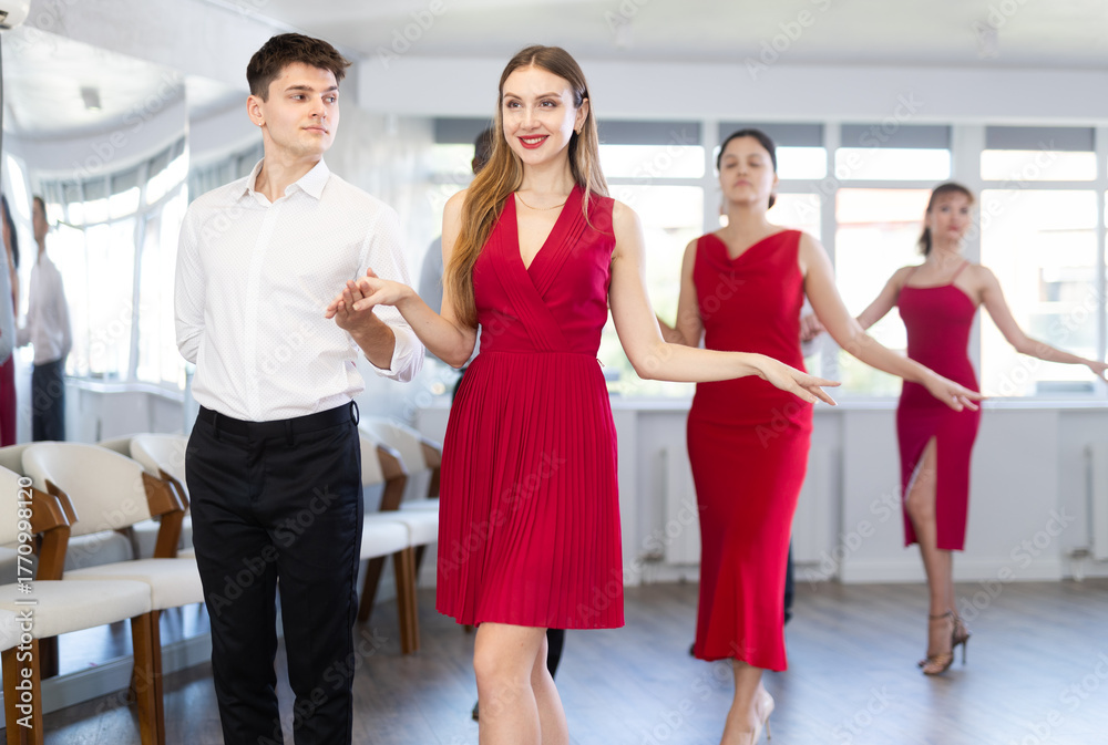 Naklejka premium Attractive elegant woman in red dress enjoying graceful waltz paired with young guy during group dance class