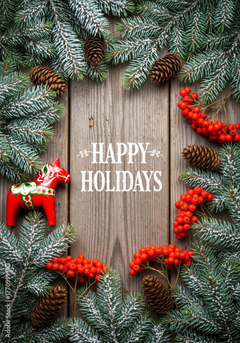 Happy Holidays Greeting With Frosted Evergreen Branches, Pinecones, Red Berries, And Wooden Background