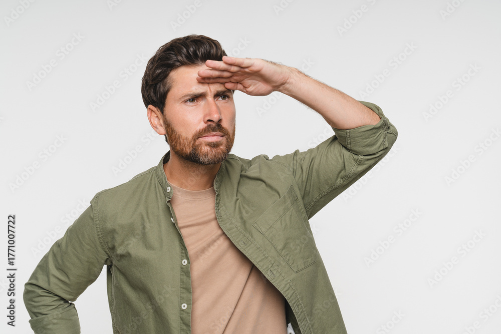 Obraz premium Portrait of man with bristle trying to see something into distance, holds palm on forehead looks away searches someone focused far away, wearing casual shirt. Indoor shot isolated on white background.