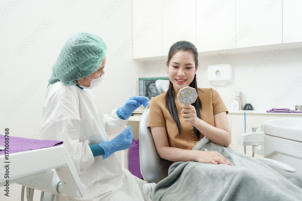 Fototapeta premium Female dental assistant in surgical attire communicating with asian patient holding mirror in dental chair, explaining teeth condition before dental procedure in modern clinical setting
