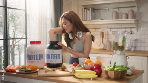 Young Asian woman in sportswear making healthy protein shake smoothie in home kitchen, using organic fresh fruits, vegetables, and whey powder. Wellness, fitness lifestyle, balanced diet, weight loss.